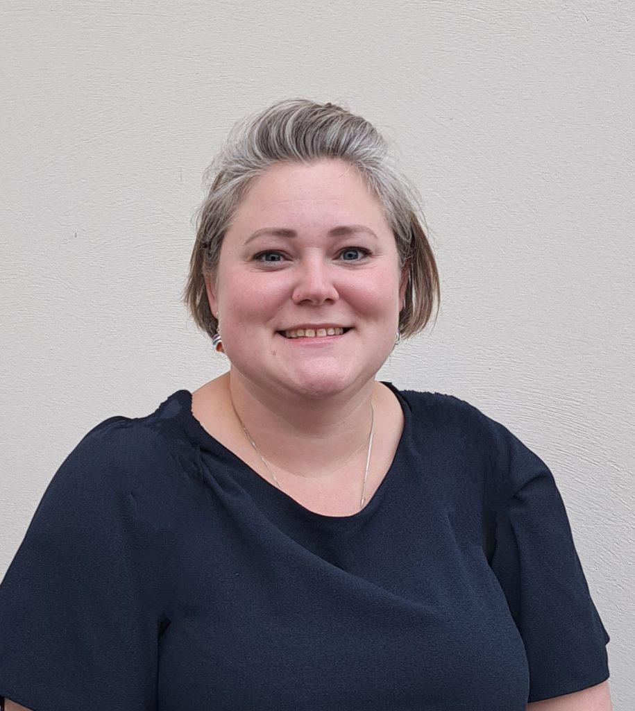 A white lady in her thirties with grey and light brown hair shaped in to a short bob. She is wearing a round neck navy blue dress
